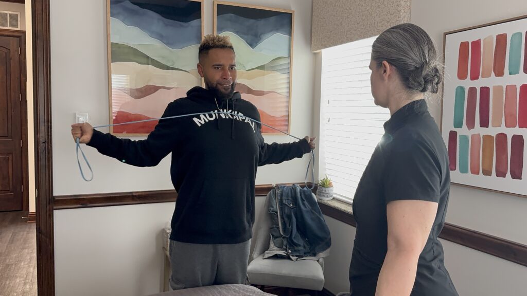A person in a black hoodie stretches a resistance band with both arms while another person observes in a room with colorful artwork on the walls.