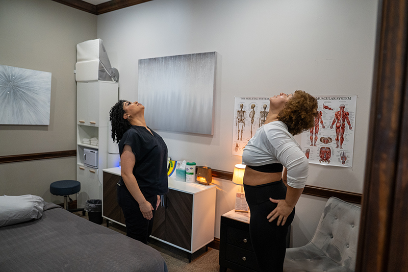 Two women stand in a medical office, both tilting their heads back, appearing to do a neck stretch; anatomical posters and medical equipment are visible in the background.