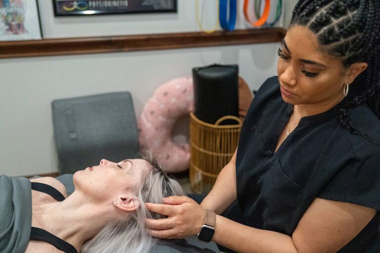 A practitioner in black scrubs gently examines the jaw and neck of a woman lying on a treatment table in a clinical setting.