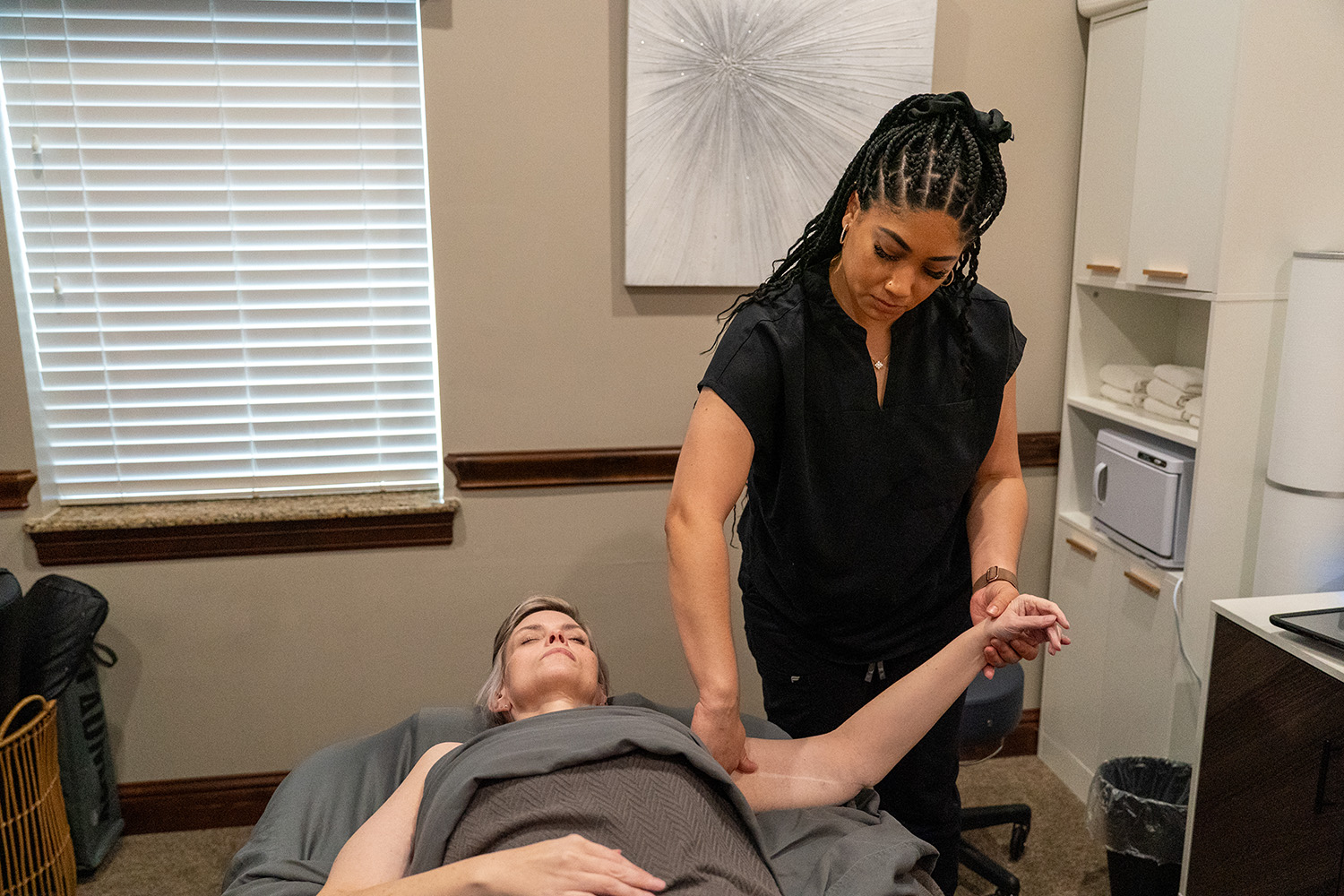 A woman lies on a massage table while a massage therapist lifts and stretches her arm in a treatment room.