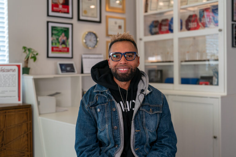 A person with glasses and a beard sits indoors, wearing a denim jacket and black hoodie. Framed certificates and awards—possibly in therapeutic massage for athletes—are displayed on the wall and shelves in the background.