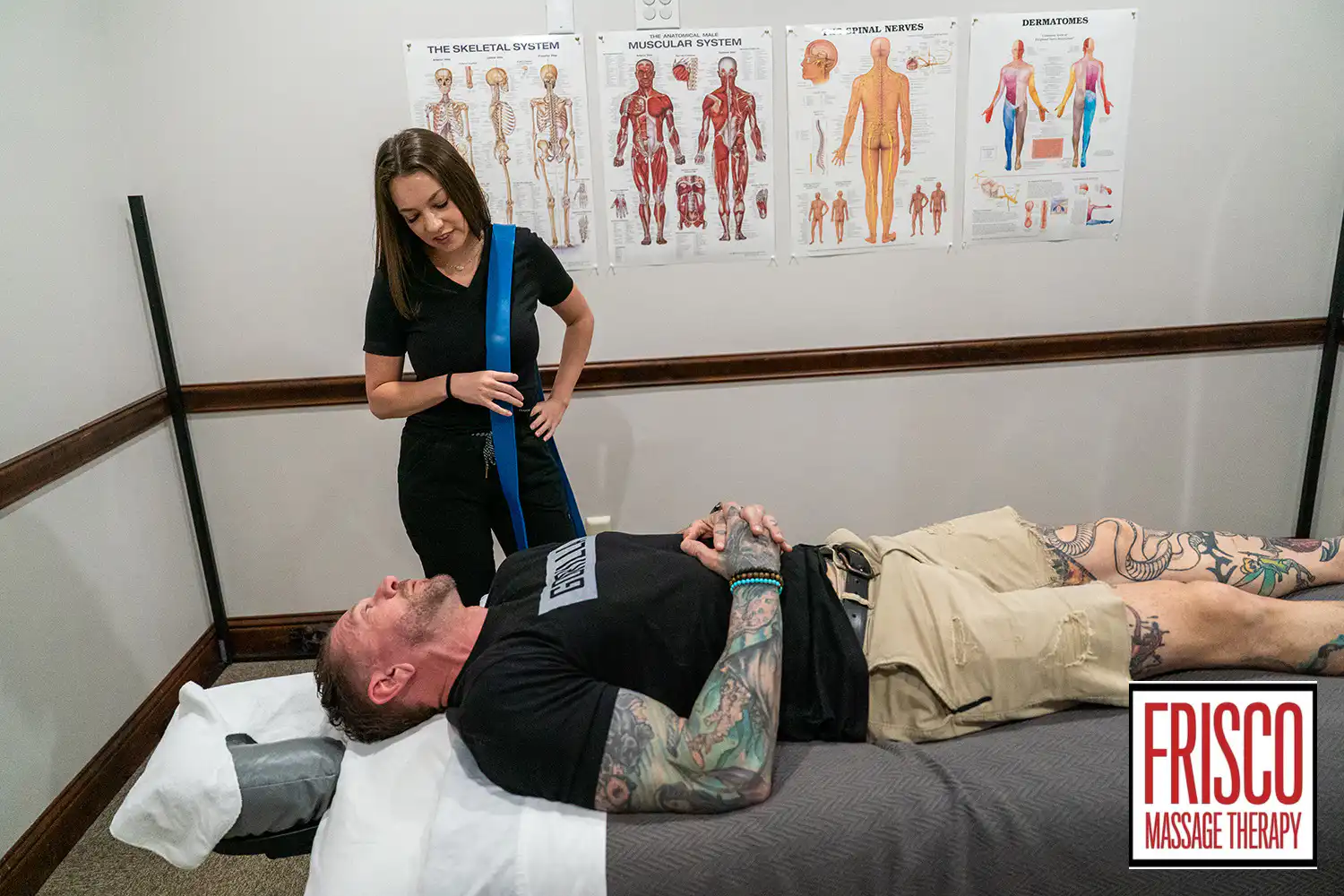 A woman stands next to a man lying on a massage table in a room with anatomical charts and cupping equipment; the Frisco Massage Therapy logo is visible.