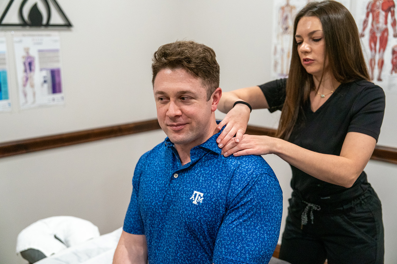 A woman in black scrubs massages the shoulders of a seated man wearing a blue Texas A&M polo in a medical or therapy office.