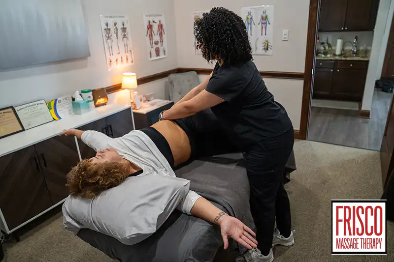 A massage therapist stretches a client's leg on a massage table in a treatment room. Anatomy charts, cupping sets, and certificates are visible on the walls. Frisco Massage Therapy logo is in the corner.