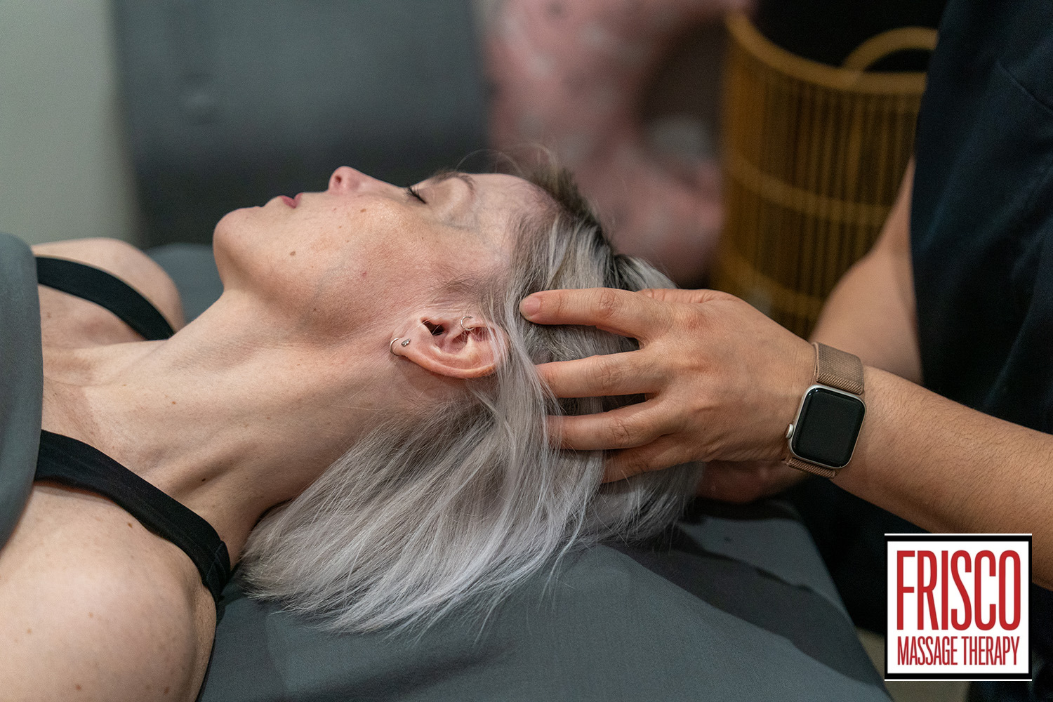 A person with light hair receives a head massage from a therapist wearing a smartwatch, specializing in manual lymphatic drainage for chronic pain; the "Frisco Massage Therapy" logo is visible in the bottom right corner.