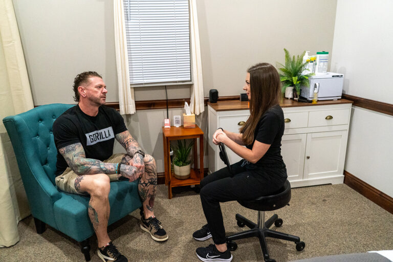 A man with tattoos sits in a teal chair speaking with a woman on a stool in a small, well-lit office with plants and medical supplies.