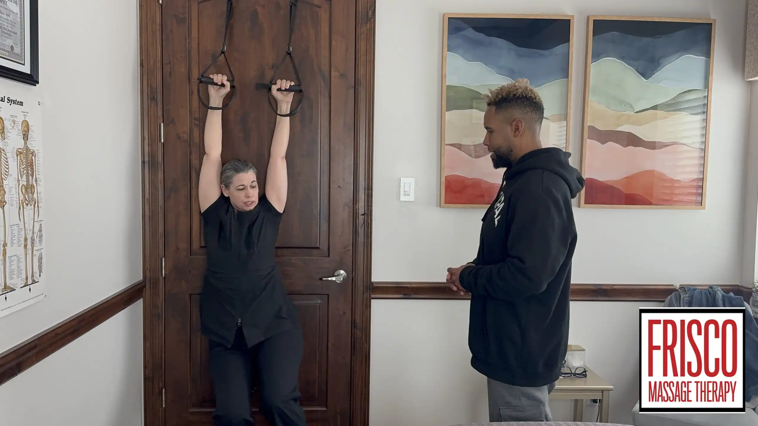 A person hangs from straps attached to a door while another observes in a room with anatomical charts, framed artwork, and hints of cupping therapy. The "Frisco Massage Therapy" logo is visible.