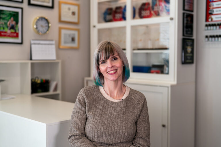 A woman with gray hair and teal highlights sits and smiles in an office setting with certificates and shelves, reflecting her expertise in lymphatic massage Frisco TX for lipedema treatment.