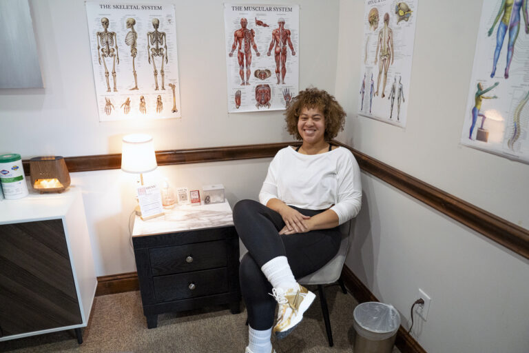 A person sits smiling in a medical office with anatomical posters of the skeletal and muscular systems on the wall behind them, perhaps awaiting a manual lymphatic drainage session.