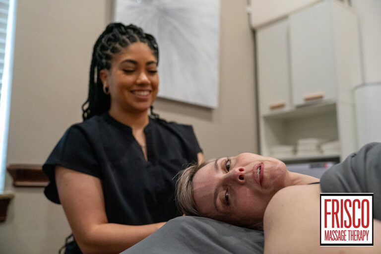 A massage therapist stands beside a client lying on a table in a treatment room, highlighting the manual lymphatic drainage benefits offered. The client looks at the camera, with the Frisco Massage Therapy logo visible in the corner.