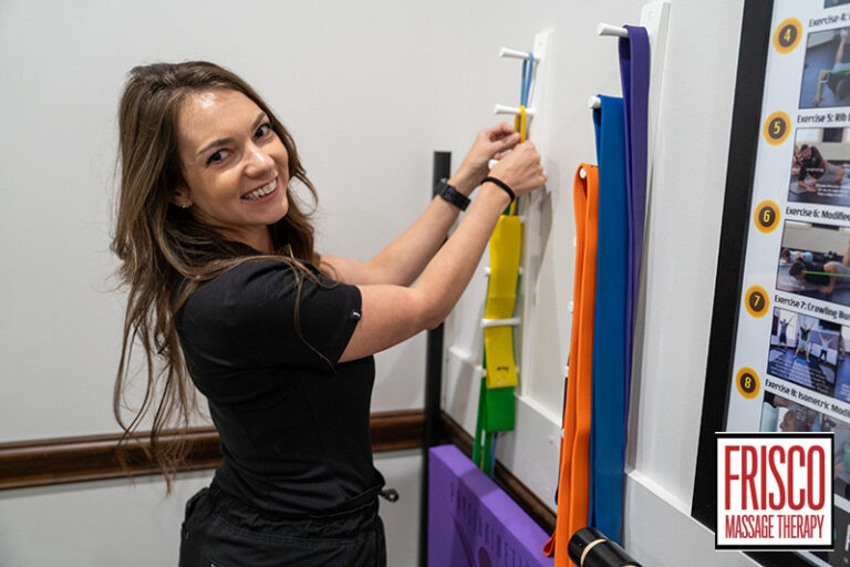 A woman smiles while organizing resistance bands on a wall rack in a clinic labeled Frisco Sports Massage Therapy.