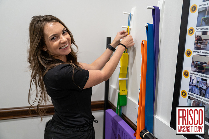 A woman smiles while organizing resistance bands on a wall rack in a clinic labeled Frisco Sports Massage Therapy.