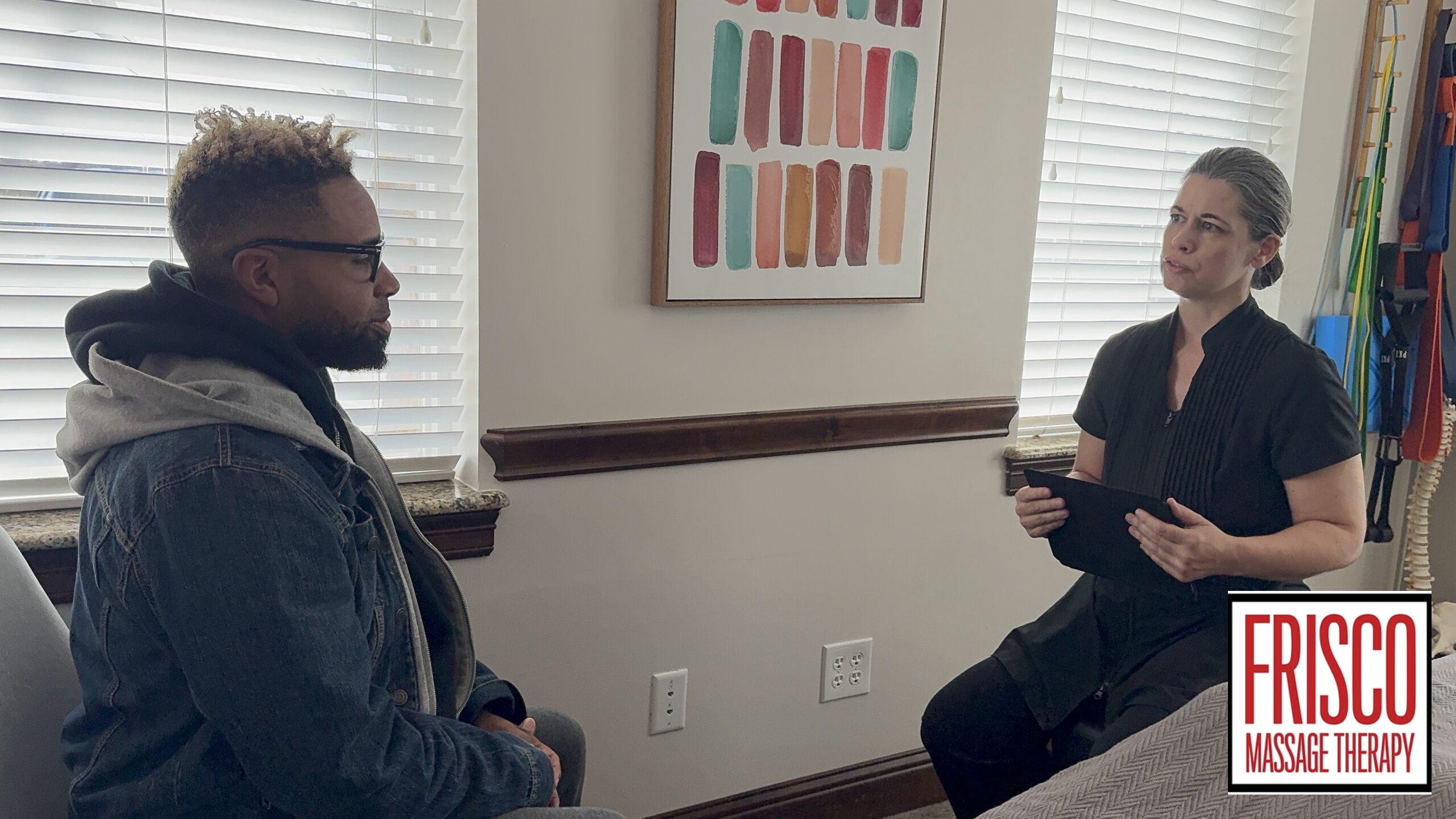 A man sits across from a woman holding a tablet in an office with blinds, wall art, and a "Frisco Massage Therapy" logo, discussing the best massage therapy for sports injuries.