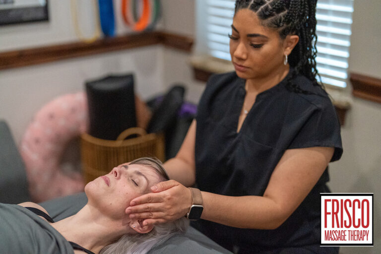 A massage therapist gently holds a client's head during a session at Frisco Massage Therapy. The client, seeking manual lymphatic drainage therapy near me, lies relaxed on the table with eyes closed as therapy tools rest in the background.