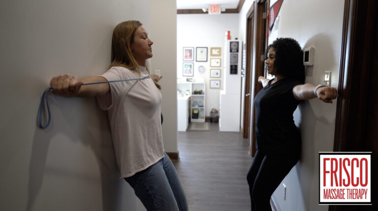Two women stand in a hallway doing a shoulder exercise with a resistance band; one instructs while the other follows at Frisco Sports Massage Therapy center.
