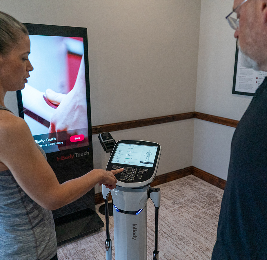A woman demonstrates a body composition analyzer to a man in a clinical setting, with a digital display screen in the background, highlighting the benefits of InBody assessment Frisco TX for comprehensive body composition analysis.