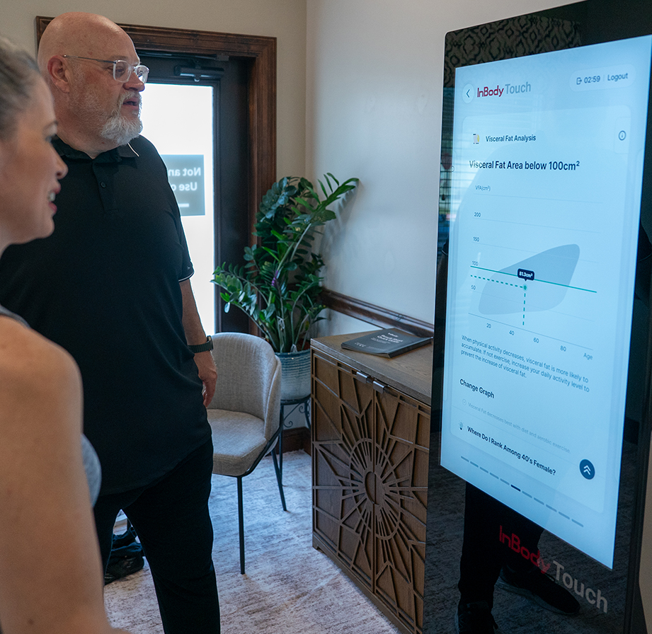 Two people stand in front of a health analysis kiosk displaying a visceral fat area chart and related data, highlighting the benefits of body composition analysis in a modern room with a plant and chairs.