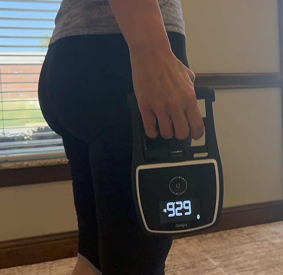Person holding a digital scale showing a weight of 62.9, standing indoors near a window, dressed in dark leggings and a gray top—an ideal setting for body composition analysis Frisco TX residents trust to track their wellness journey.