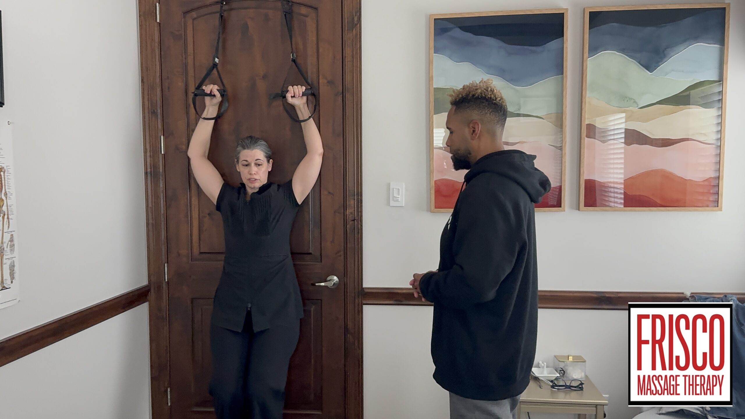 A woman practices an exercise using suspension straps on a door while a man observes, demonstrating athlete care. Abstract paintings and a Frisco Massage Therapy sign on the wall highlight the setting’s focus on wellness and therapeutic massage.