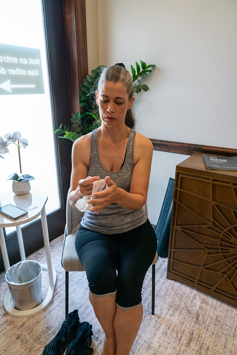 A woman in athletic wear sits on a chair, wiping her hands with a towel in a room with a plant, a small table, and a sign on the glass door.