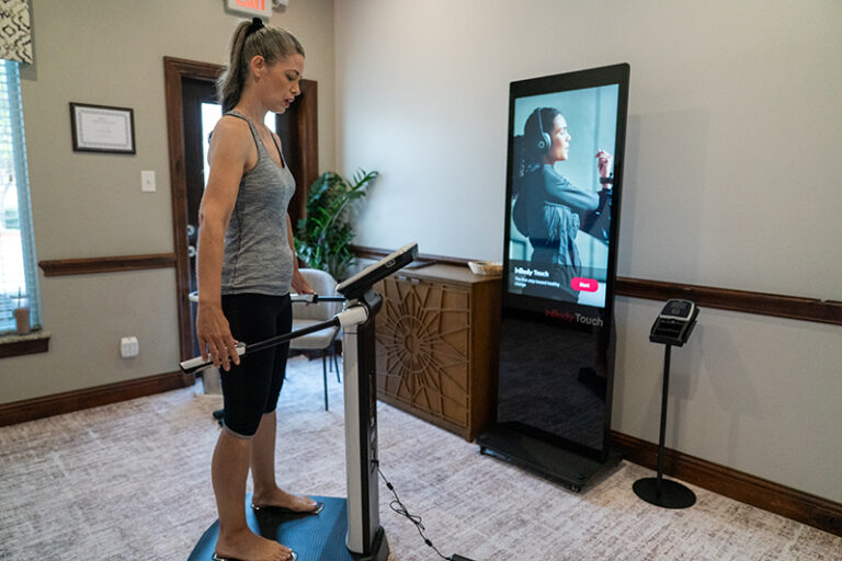 Woman stands on a fitness platform holding handlebars, facing a screen displaying her image during a health assessment in a modern room.