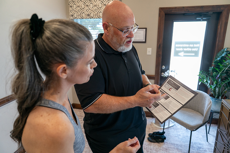 A man and a woman stand in an office, reviewing information on a brochure together. The man is pointing at the brochure while the woman attentively listens.