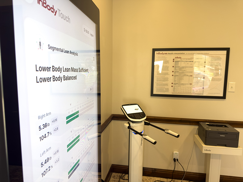 A body composition analysis machine and monitor displaying test results in a medical office, with a printer and framed chart on the wall.
