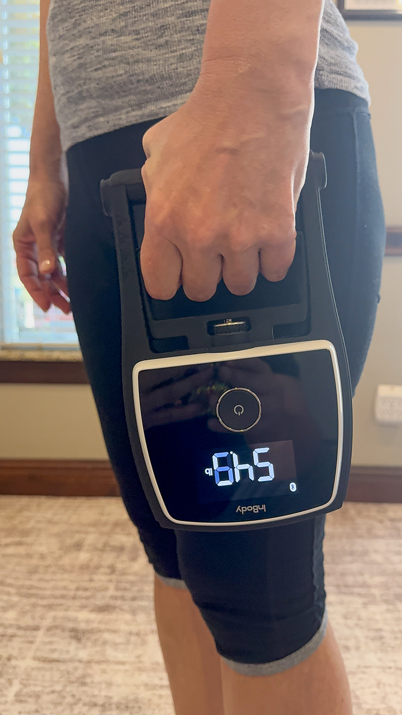 A person in exercise clothes holds a digital scale displaying a weight of 6.45 pounds, with their arm extended by their side indoors.