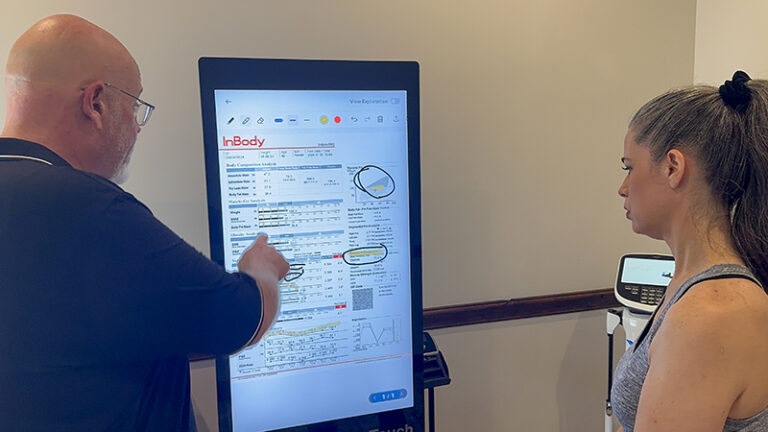 A man points at a digital screen displaying health data while a woman looks on, both standing indoors near health monitoring equipment.