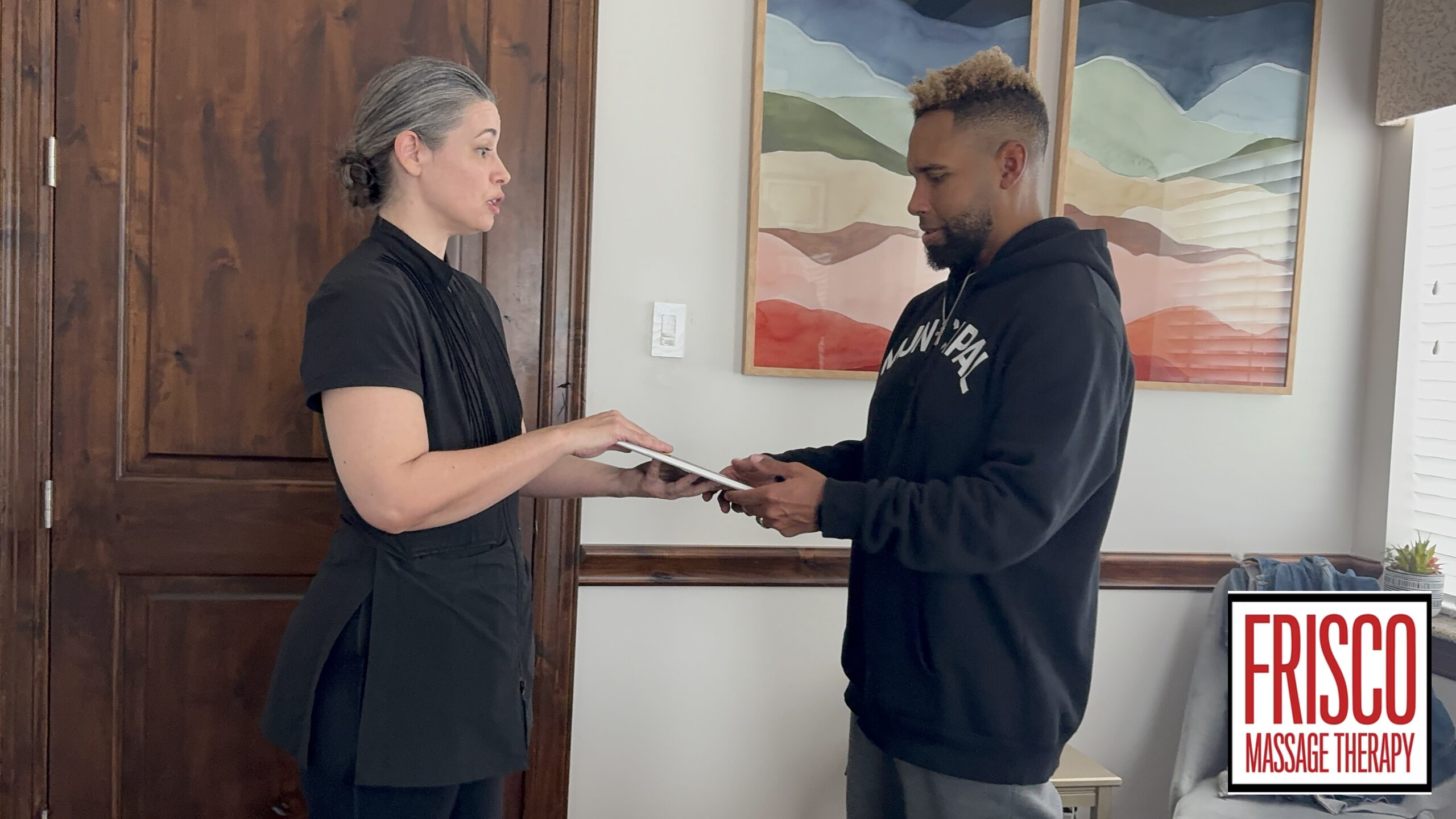 A woman hands paperwork to a man in a casual room, suggesting an intake process for Frisco sports massage therapy. The Frisco Massage Therapy logo is visible in the lower right corner.