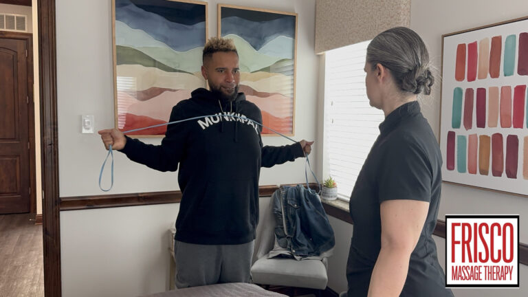 A man in a black hoodie performs a resistance band exercise guided by a massage therapist in a treatment room at Frisco Massage Therapy, known for providing the best massage therapy for sports injuries.