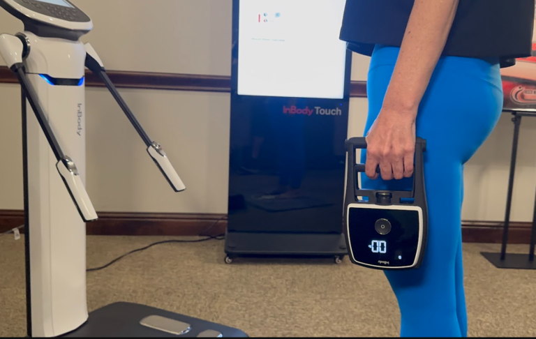 Person in blue pants holding a handheld device near a body composition scanner in a room with medical equipment and display screens for grip strength and cognitive health.