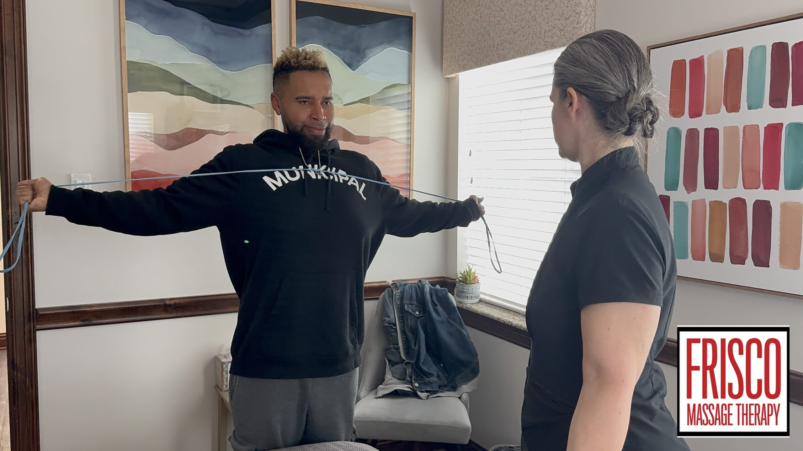 A person stands with arms outstretched holding a resistance band, facing another person in an office with the “Frisco Massage Therapy” logo in the corner, highlighting their reputation for offering the best massage therapy for sports injuries.
