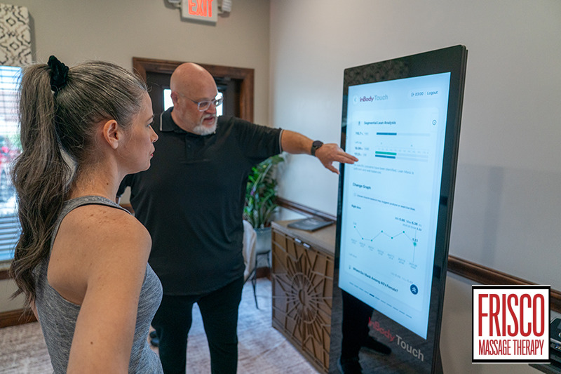 A man points at data on a large touchscreen, explaining the results of an InBody 580 body composition analysis to a woman in an indoor setting. The “Frisco Massage Therapy” logo is visible in the bottom right corner.