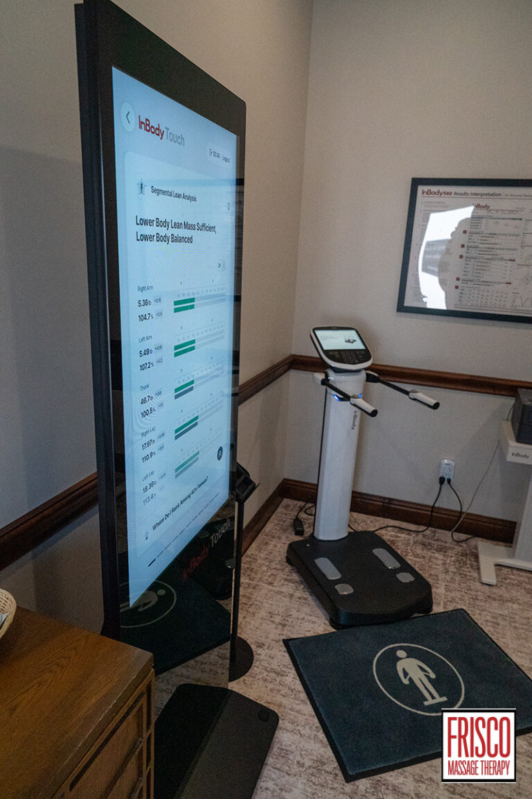 A digital body composition analyzer, designed for precise body composition analysis, features a touchscreen that displays results next to a body scale and mat in a massage therapy clinic. The Frisco Massage Therapy logo is visible in the corner.