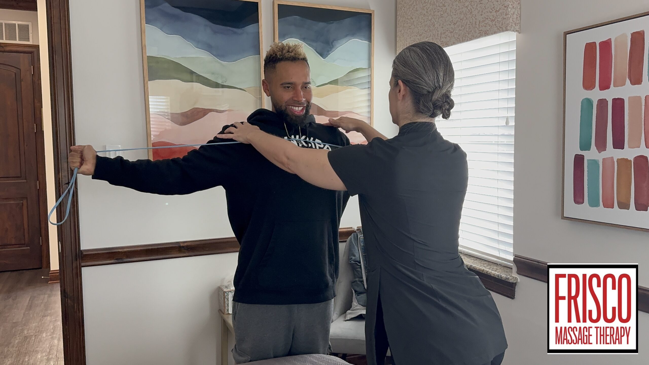 A massage therapist measures a client’s arm span with a tape measure in a well-lit room, demonstrating how therapeutic massage aids in post-surgery recovery. The Frisco Massage Therapy logo is visible in the corner.