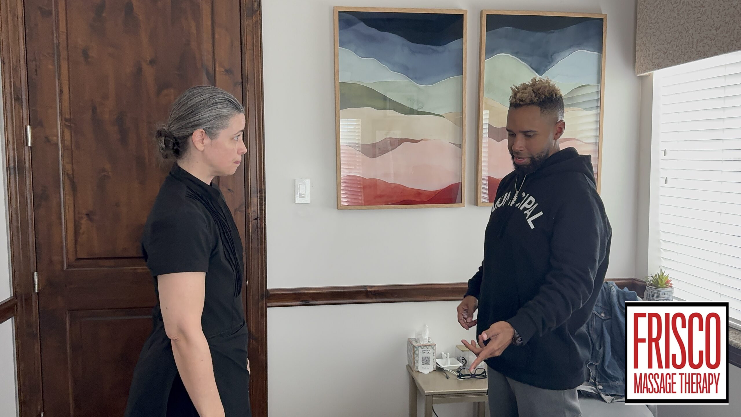A woman and a man stand in a therapy room with abstract art and a small table, highlighting the serene environment ideal for sports therapy massage in Frisco. The Frisco Massage Therapy logo is visible in the bottom right corner.