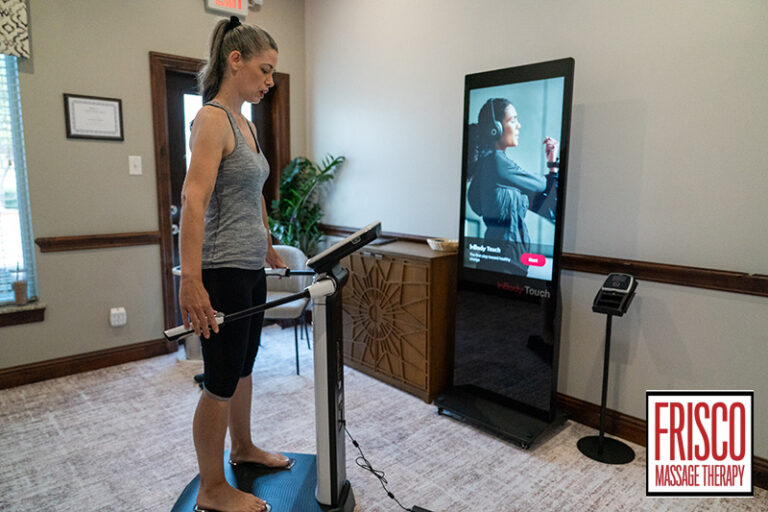 A woman stands on an InBody 580 body composition analyzer in a clinic room, with a fitness screen and Frisco Massage Therapy logo visible, highlighting the advanced body composition analysis services available in Frisco.