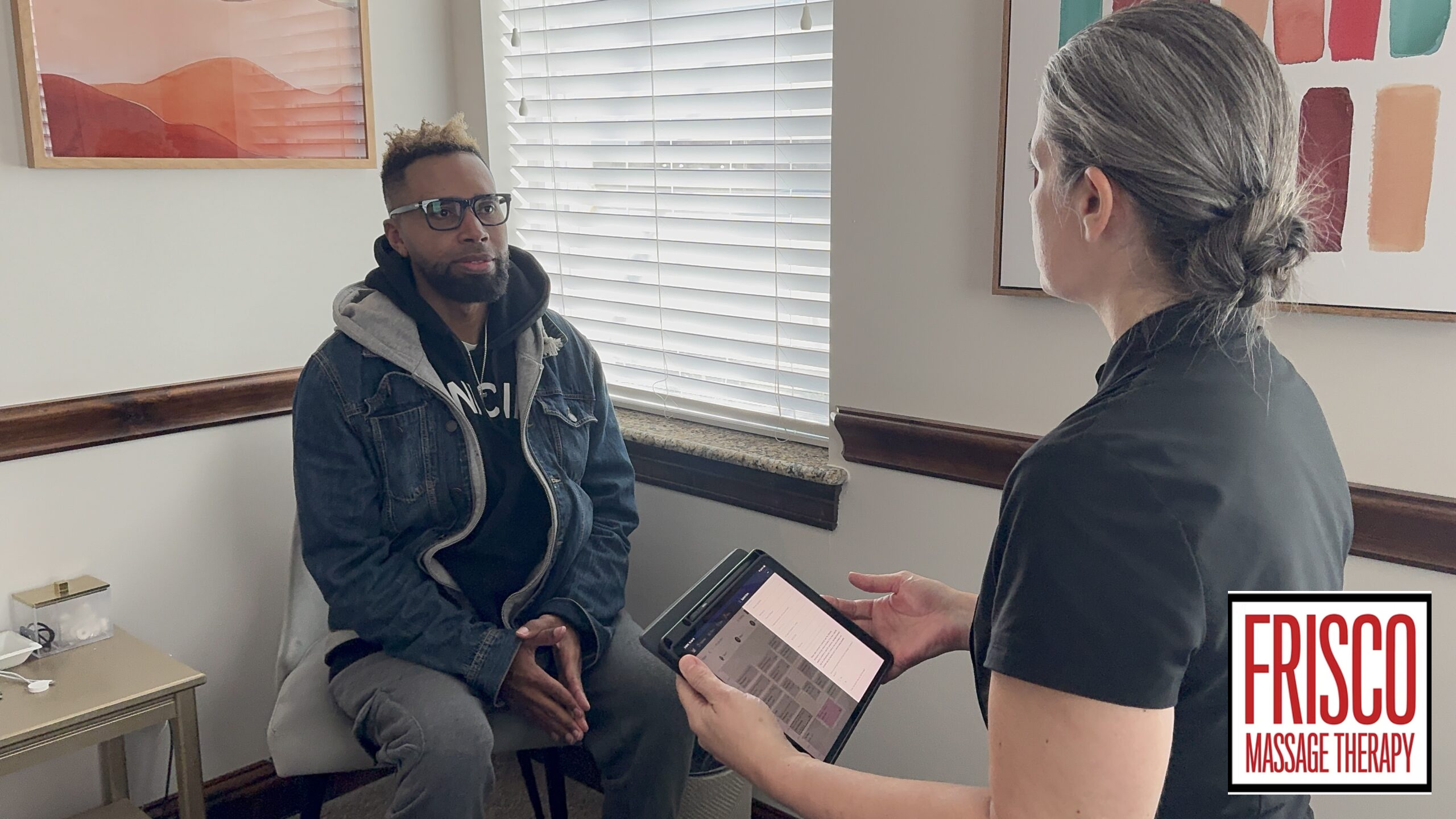A massage therapist shows a client a digital chart on a tablet to explain how therapeutic massage aids in post-surgery recovery.