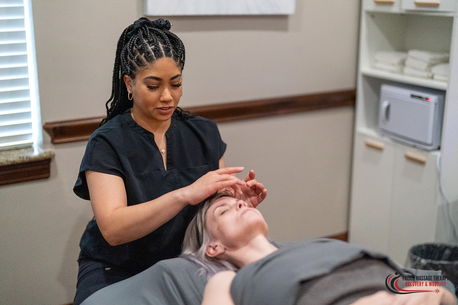 A massage therapist performing a scalp and facial massage for lymphatic drainage on a client lying on a treatment table.
