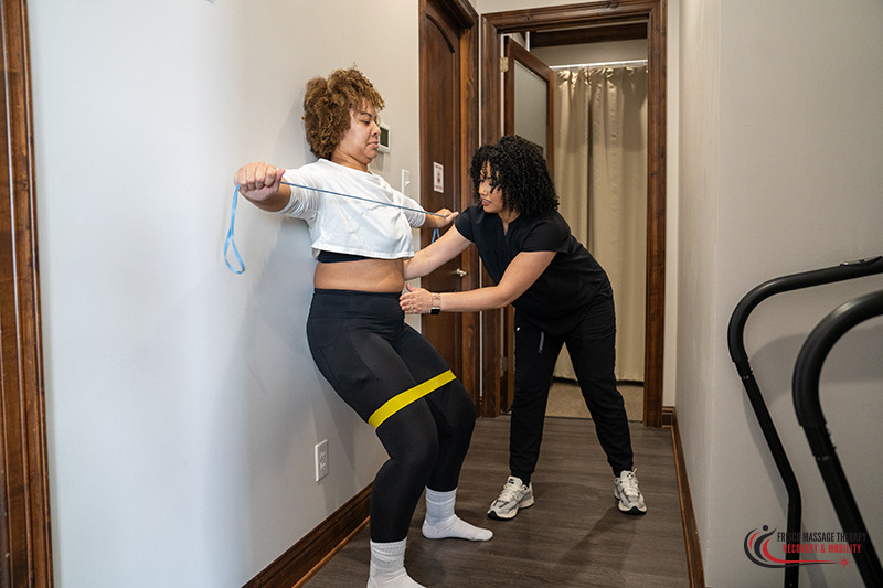 A physical therapist supervises a client performing a wall sit with a resistance band and a chest stretch during a sports therapy massage session in Frisco.