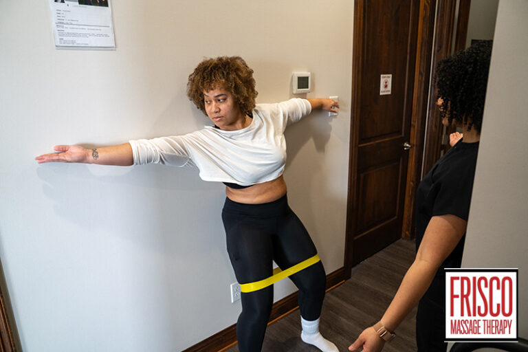 A woman with a yellow resistance band around her thighs leans against a wall to perform a shoulder stretch while a sports therapy massage therapist in Frisco observes.