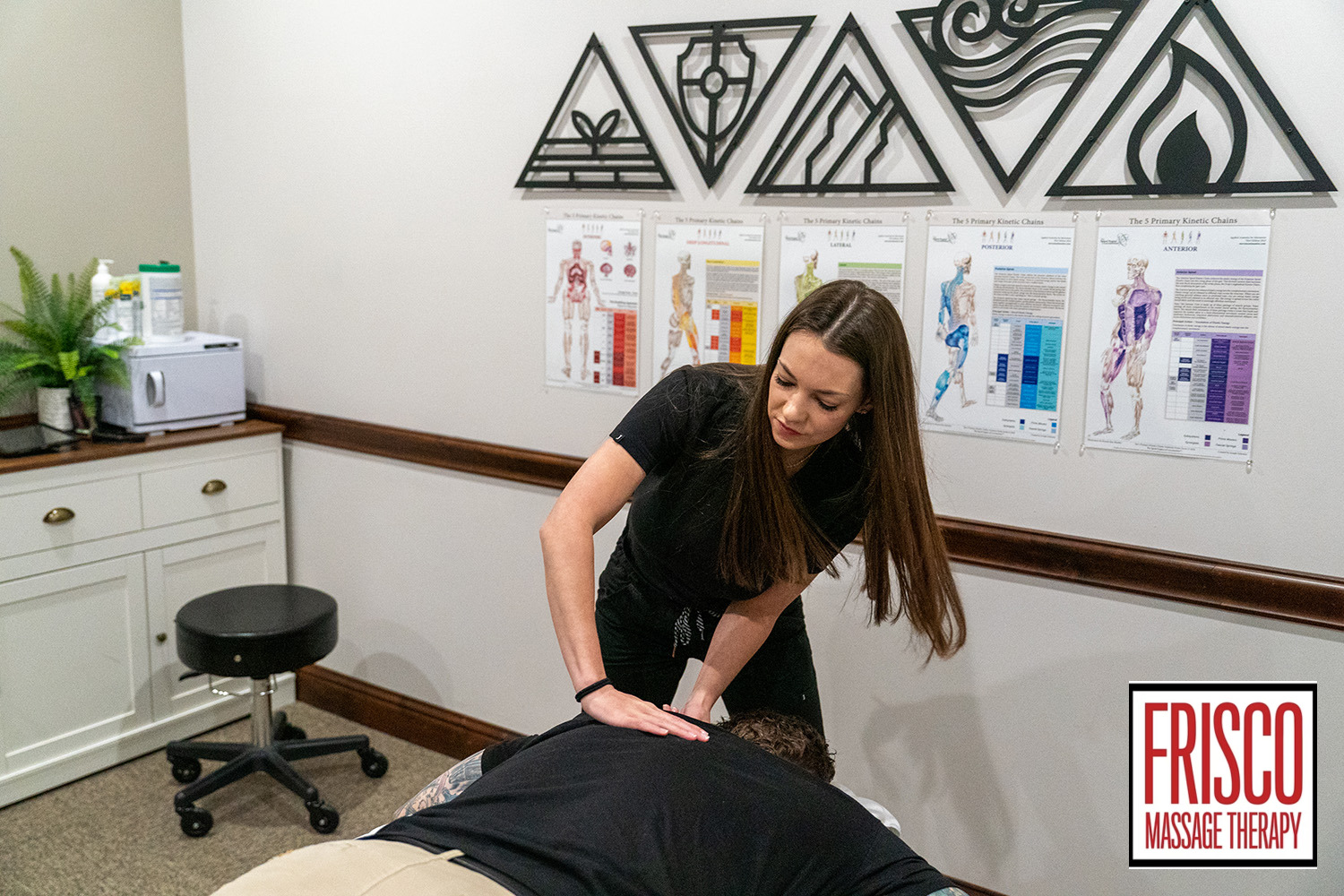 A massage therapist performing manual therapy on a client’s back in a clinic decorated with anatomical charts, demonstrating how therapeutic massage aids in post-surgery recovery.